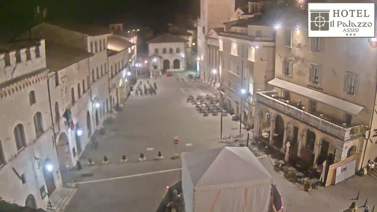 A nighttime, high-angle view of a town square with several buildings, some outdoor seating, and a temporary structure.