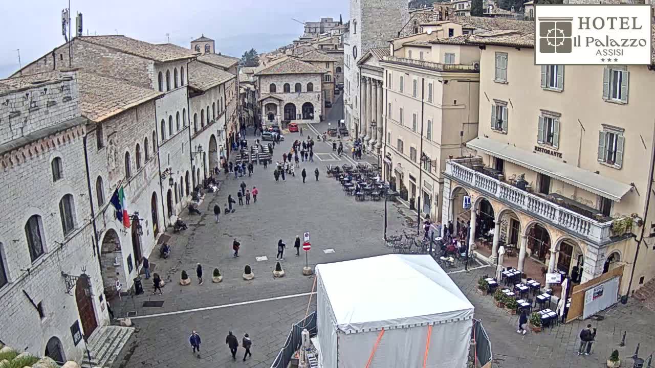A mostly empty town square in overcast weather is surrounded by historical buildings, with several people walking around and some outdoor seating areas.