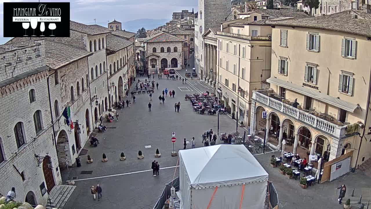 An overcast day reveals a large town square surrounded by historic stone buildings, with people scattered about and several outdoor cafes set up.