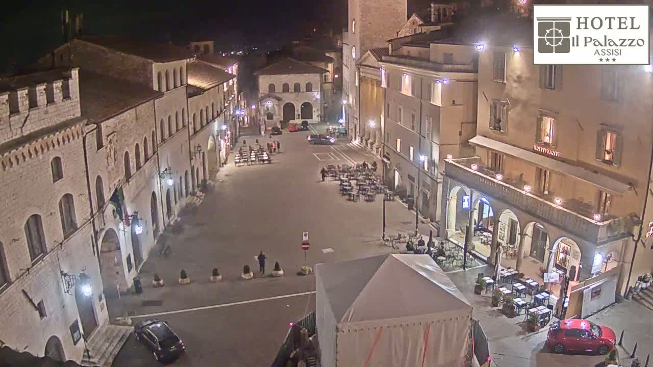 A nighttime view of a town square in Italy, with several buildings illuminated, cars parked, and outdoor seating at a restaurant.
