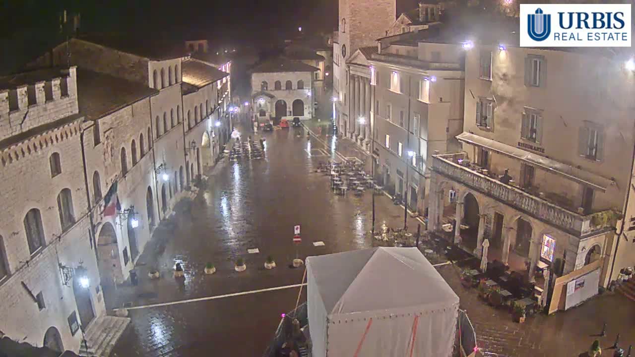 A rain-soaked town square at night, surrounded by historic buildings with outdoor seating areas, and a large tent in the center.