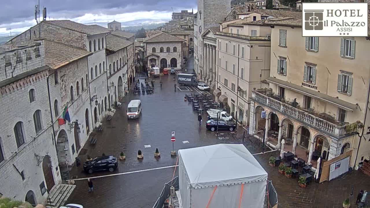 A wet, stone town square, surrounded by old buildings, features several cars, a large white tent, and outdoor seating areas under an overcast sky.