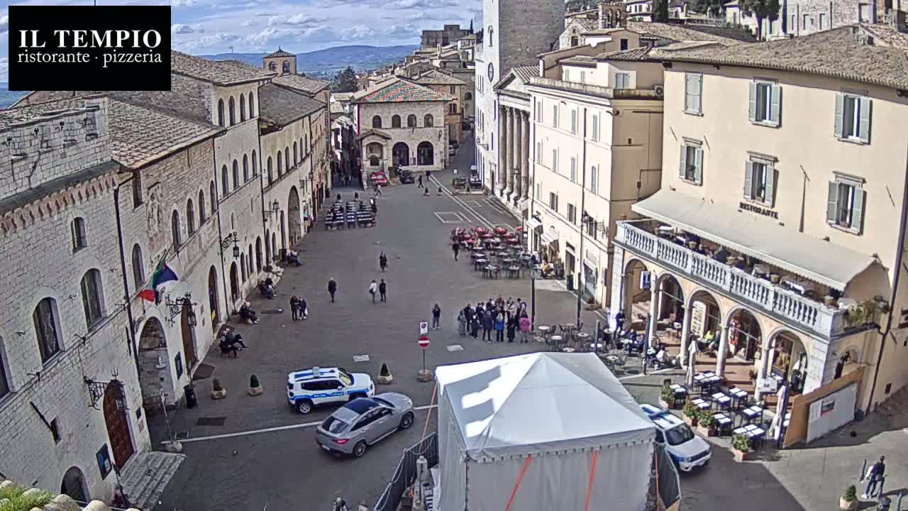 A mostly sunny, outdoor view of a large town square with several buildings, outdoor seating areas, and a few vehicles, including police cars.