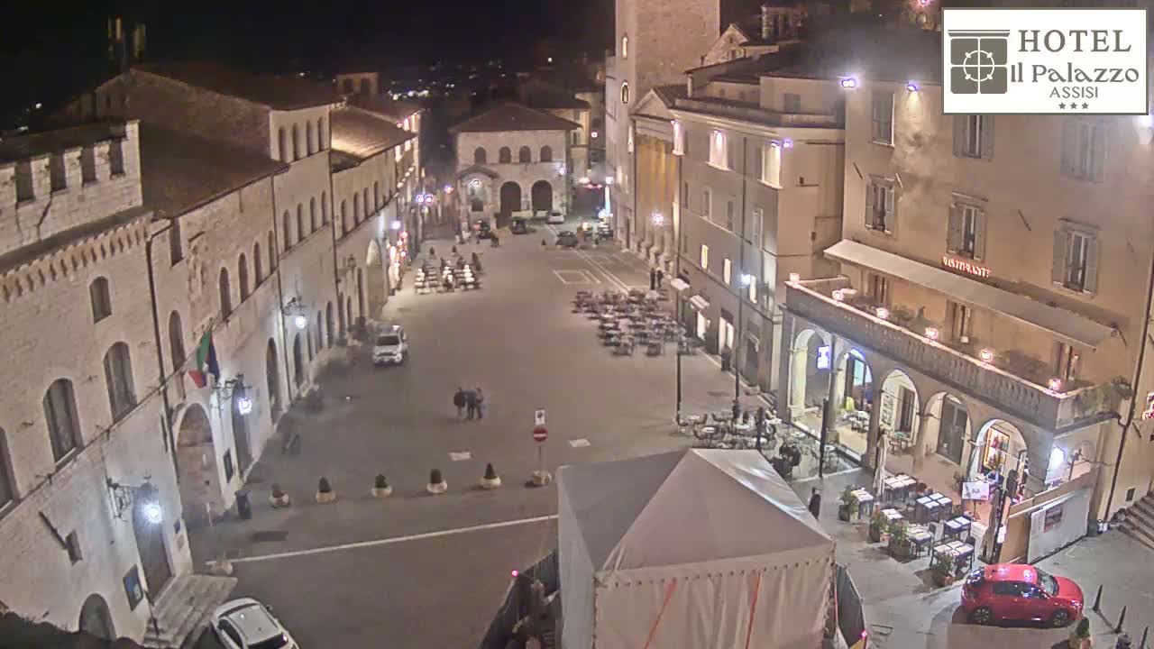A nighttime, high-angle view of a town square in Italy, showing several buildings with outdoor seating and few pedestrians.