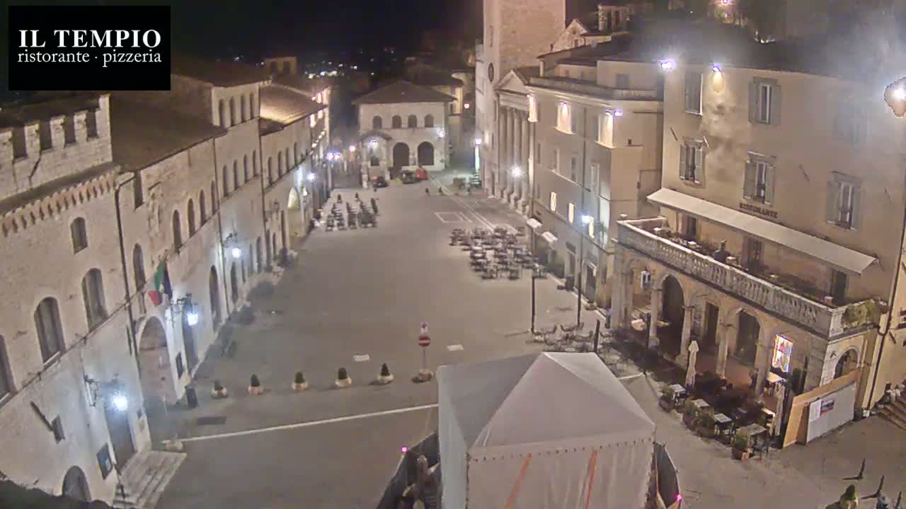 A nighttime view of a town square with several buildings surrounding it, outdoor seating at restaurants, and a large tent in the foreground.