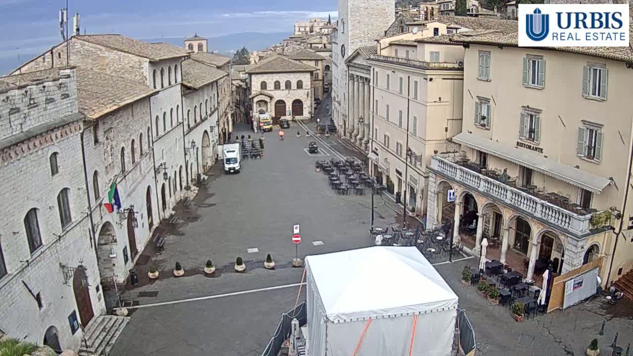 A mostly empty town square in an overcast day, surrounded by old stone buildings, with a few people and outdoor seating areas, and a large white tent in the center.