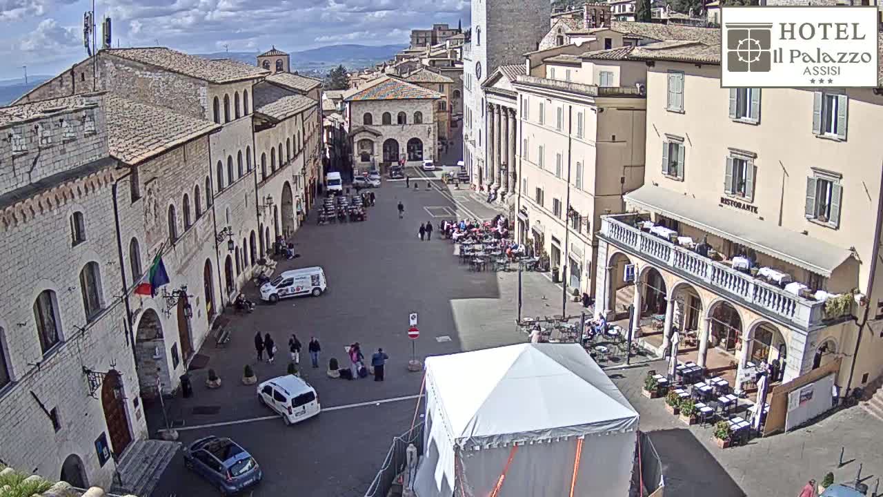 A mostly sunny town square is populated with people sitting at outdoor cafes and walking around, with several vehicles parked nearby and a large white tent in the center.