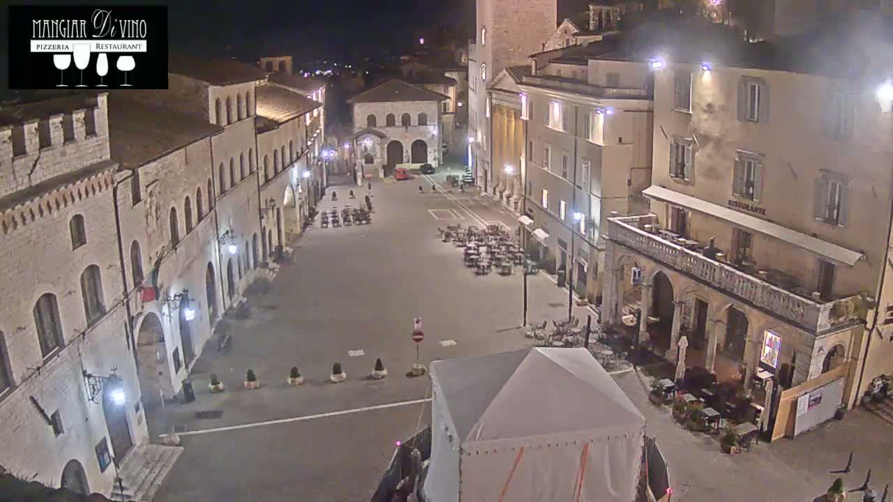 A nighttime view of a town square, illuminated by streetlights, with several buildings surrounding it and outdoor seating areas at some of the establishments.