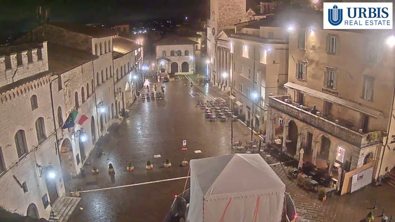 A wet, nighttime view of a town square surrounded by historic buildings, with some outdoor seating and a large tent in the center.
