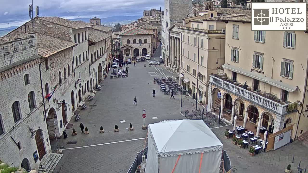 A large, empty town square with stone buildings on all sides, under an overcast sky, contains several sparsely occupied outdoor cafes and a large white tent in the center.