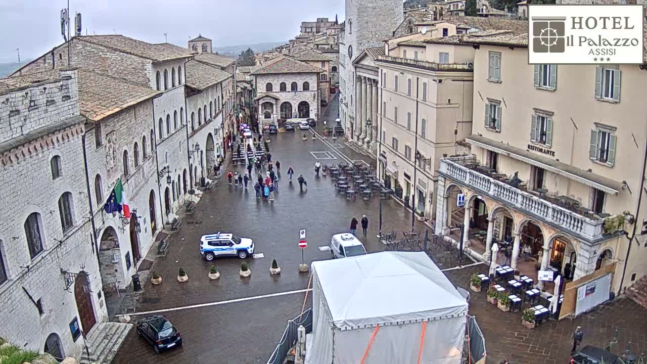 A wet town square in a historic European town is surrounded by stone buildings, with a few people and several cars visible.
