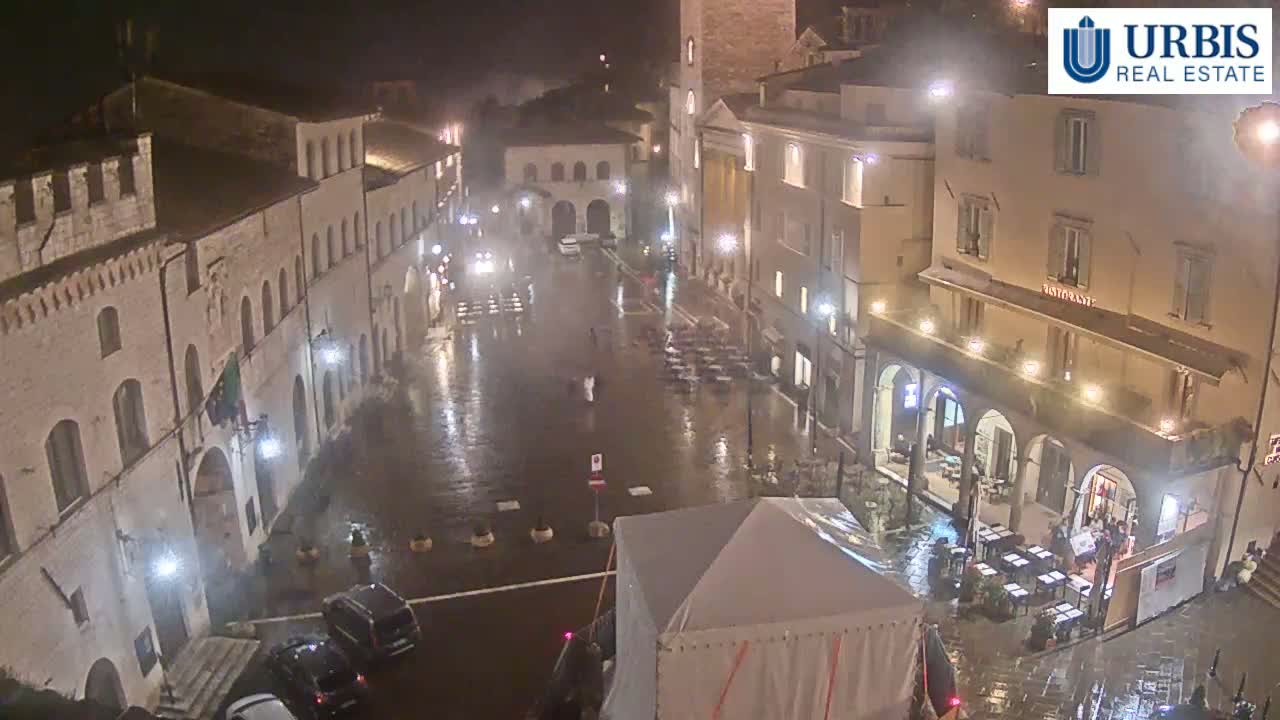 A rain-slicked town square at night is surrounded by buildings with arched doorways and outdoor seating at a restaurant.