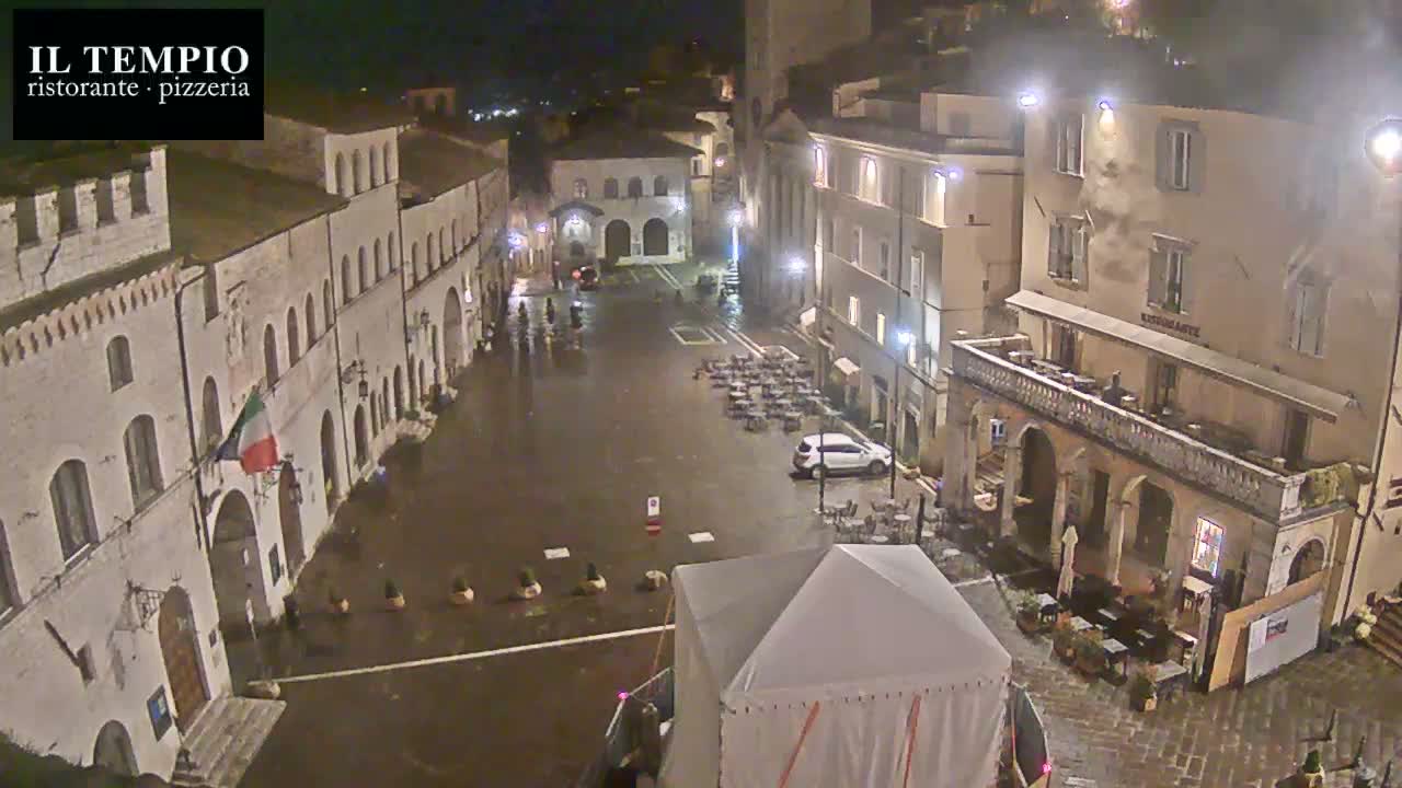 A wet, nighttime town square is surrounded by historic buildings, with outdoor seating areas and a large tent in the center.