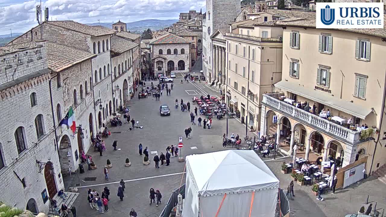 A town square in Italy, under partly cloudy skies, is filled with people sitting at outdoor cafes and walking around; a large white tent sits in the center of the square.