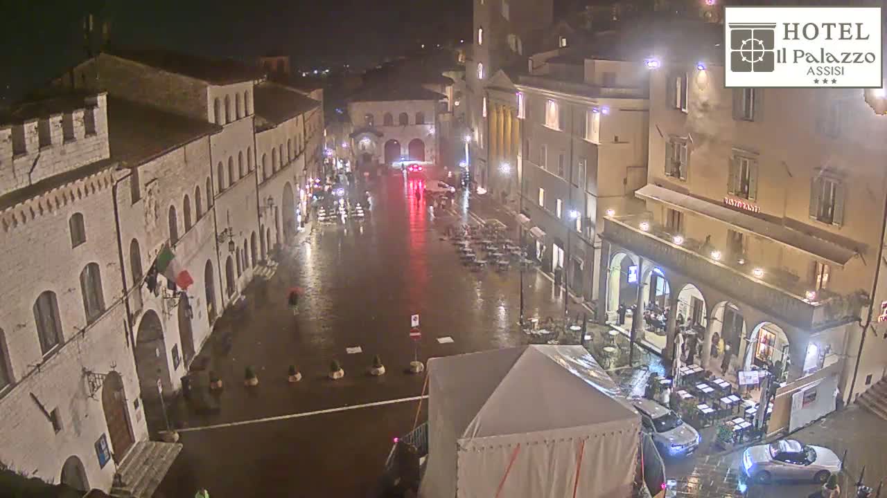 A rain-slicked town square at night is surrounded by illuminated buildings, with a few cars and people visible.