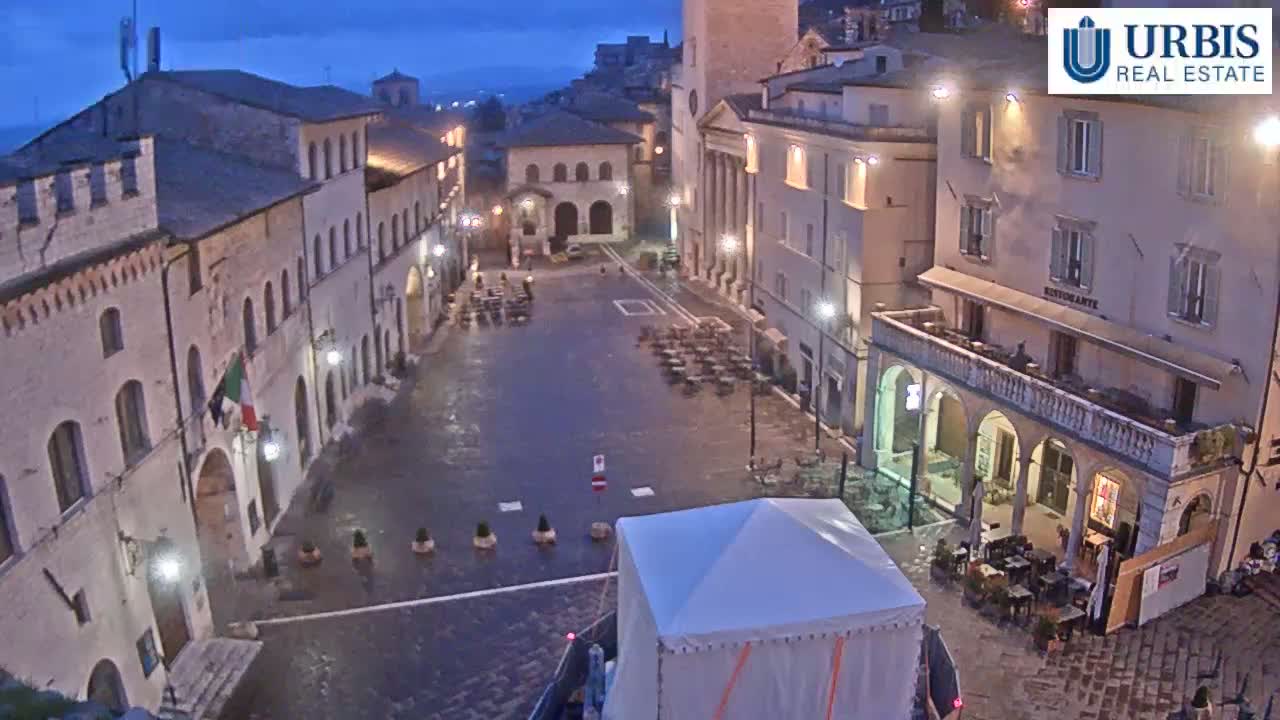 A mostly empty town square at twilight, surrounded by historic buildings, is lightly sprinkled with rain.