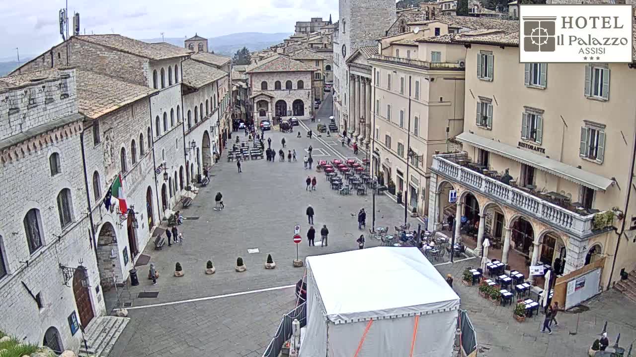 A large, paved town square is populated with a few people, outdoor seating areas, and buildings of varying heights, all under an overcast sky.
