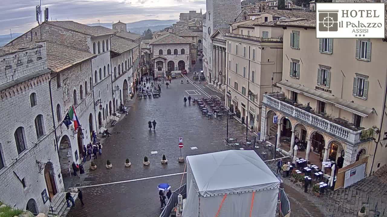 A wet, overcast town square is surrounded by several stone buildings, with some people walking about and outdoor seating areas set up.