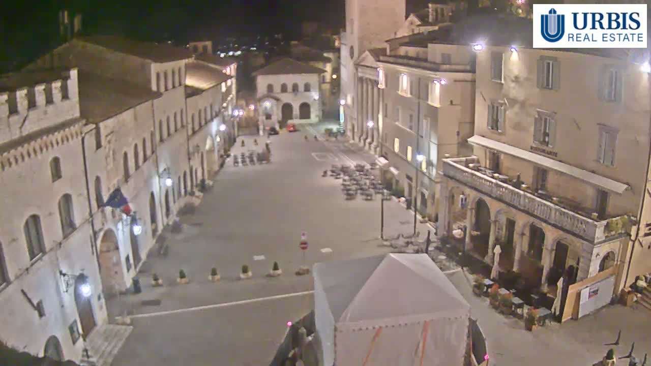 A nighttime view of a town square surrounded by historical-looking buildings, with some outdoor seating and a covered structure in the center.