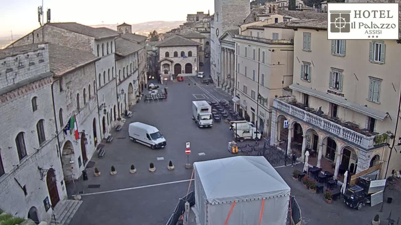 A mostly empty town square in the early morning, surrounded by stone buildings, with several vans parked in the center.