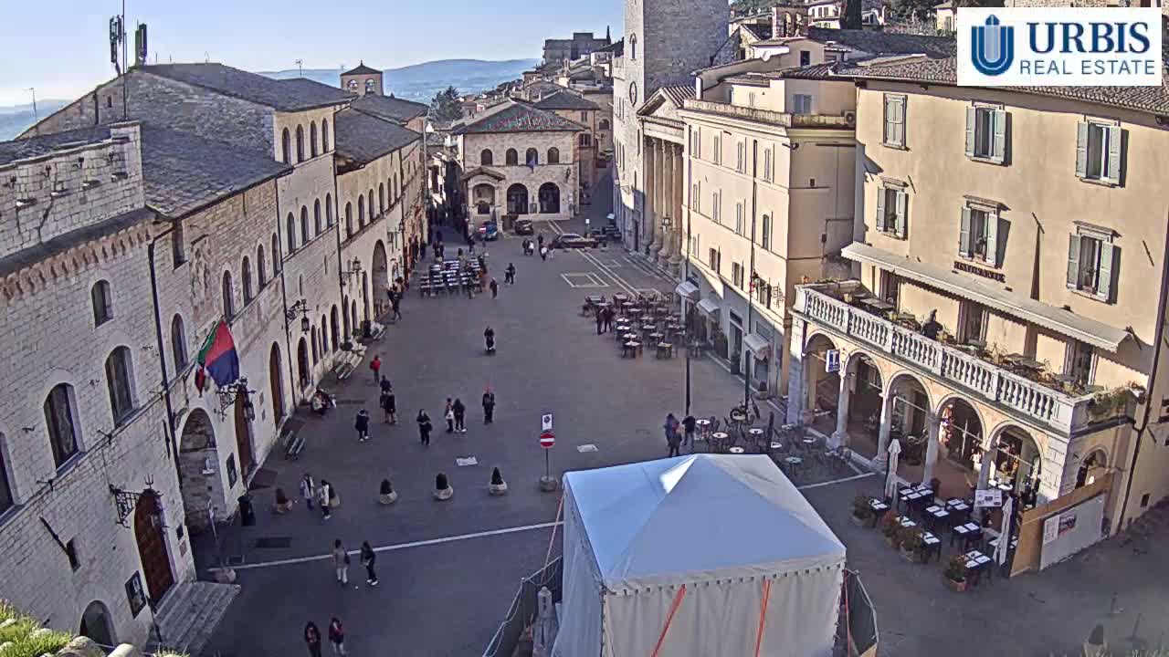 A town square in a historic European town on a sunny day is filled with people and outdoor seating at cafes, with buildings lining the perimeter.