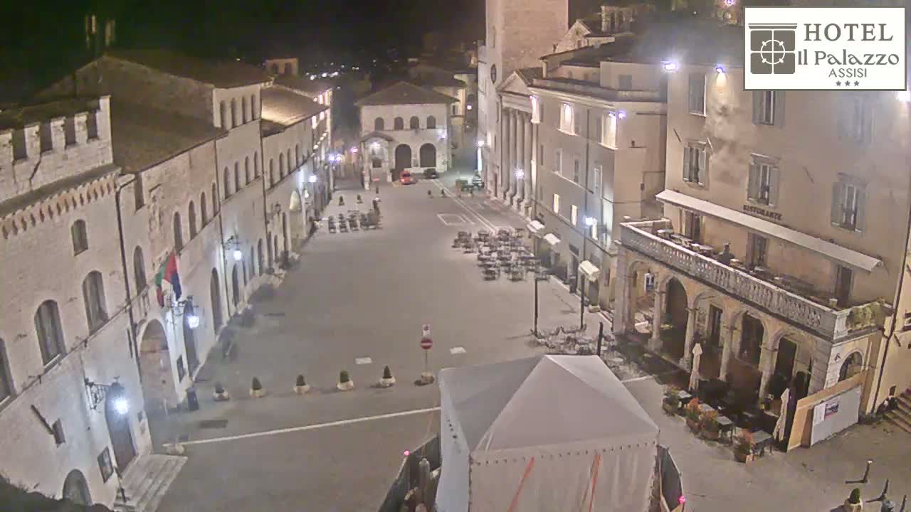 A nighttime, high-angle view of a town square with several buildings, outdoor seating areas, and a large tent in the foreground.