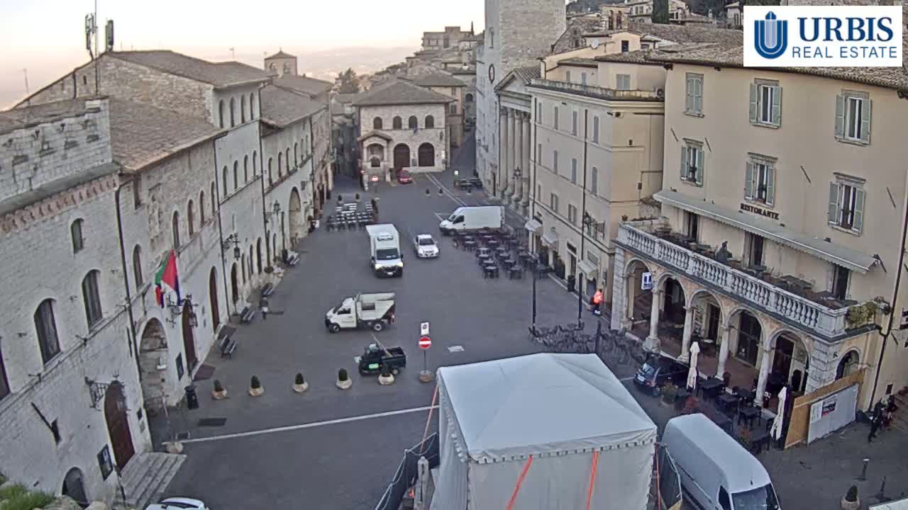 A large, mostly empty town square is surrounded by light-colored buildings, with several vehicles parked and a large white tent in the center, under a bright, possibly sunrise or sunset sky.