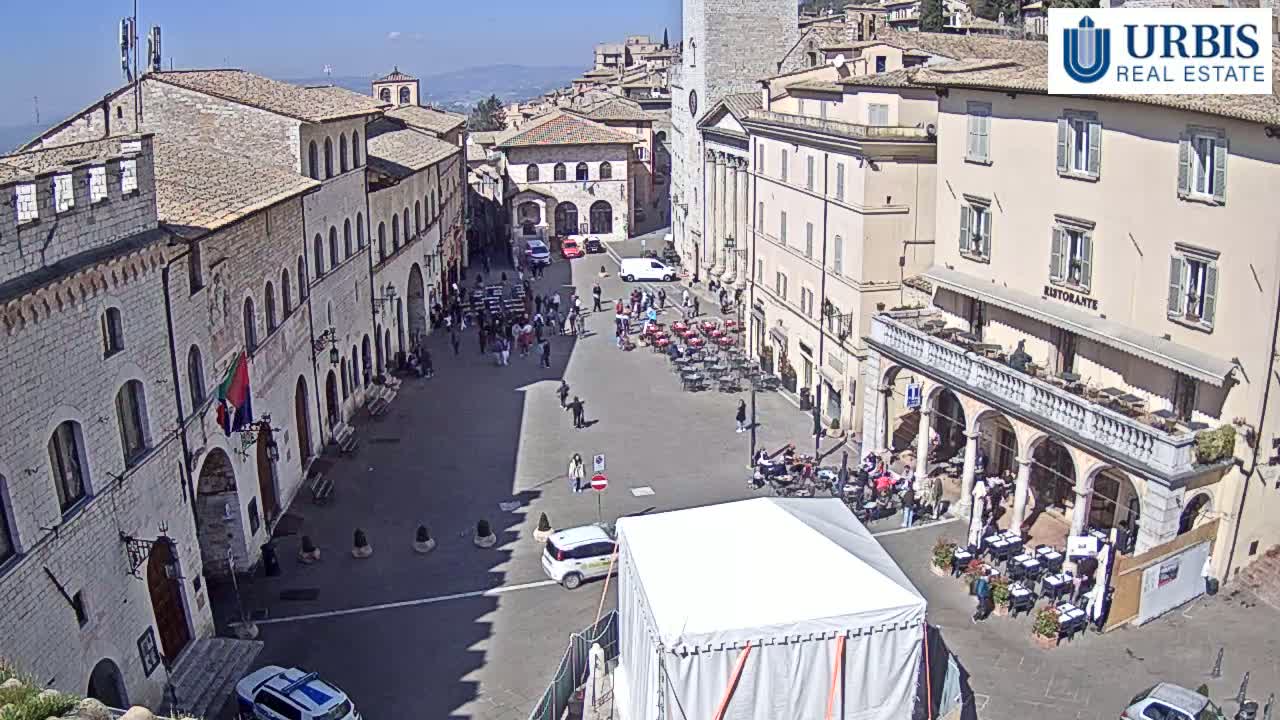 A sunny day in a European town square, showing several buildings, a few vehicles, and many people sitting at outdoor cafes and walking around.
