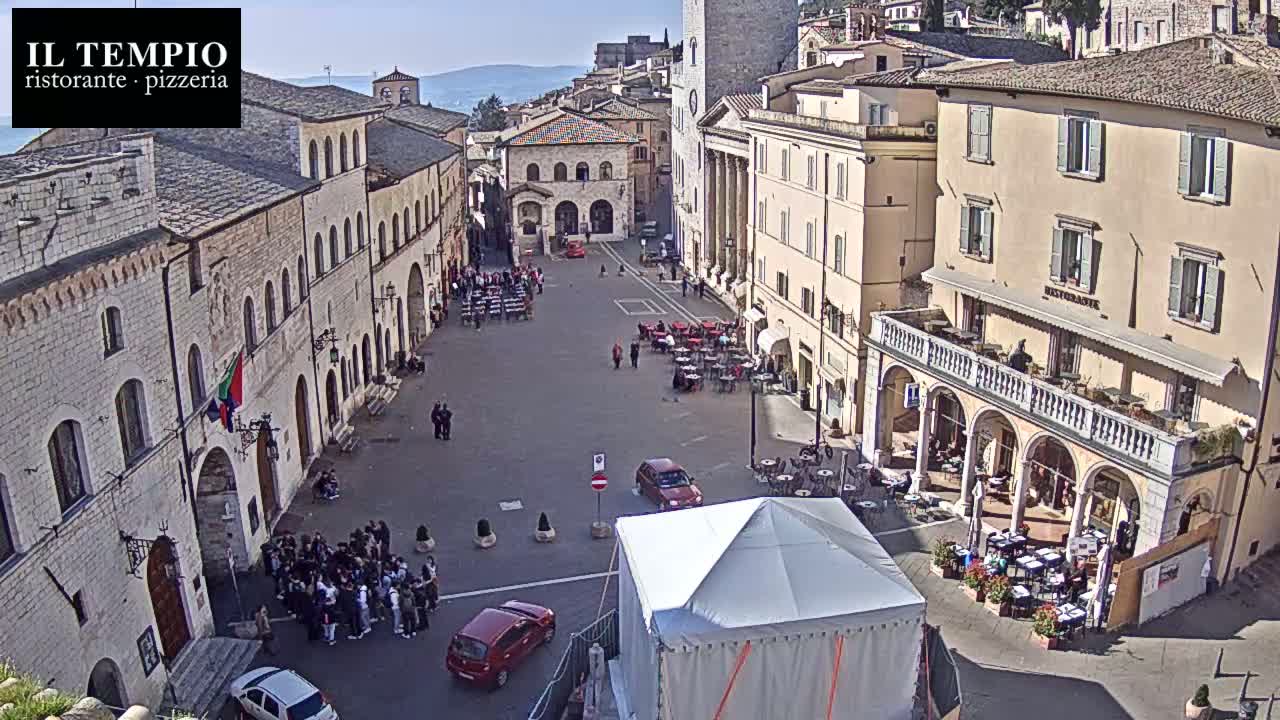 A sunny town square is filled with people, cars, and outdoor seating areas surrounding buildings of varying heights and architectural styles.