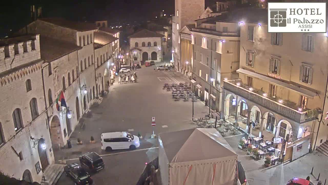 An empty, stone town square at night is illuminated, surrounded by buildings with outdoor seating at restaurants.