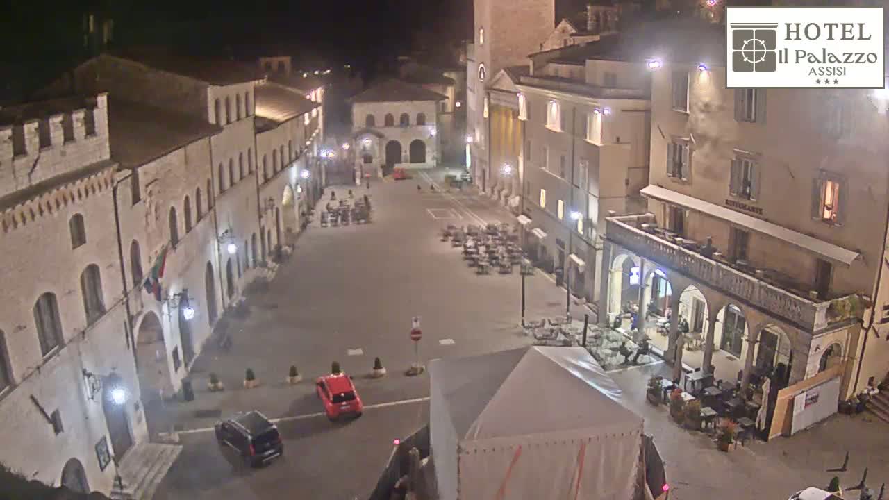 A nighttime, high-angle view of a sparsely populated town square surrounded by historic-looking buildings.