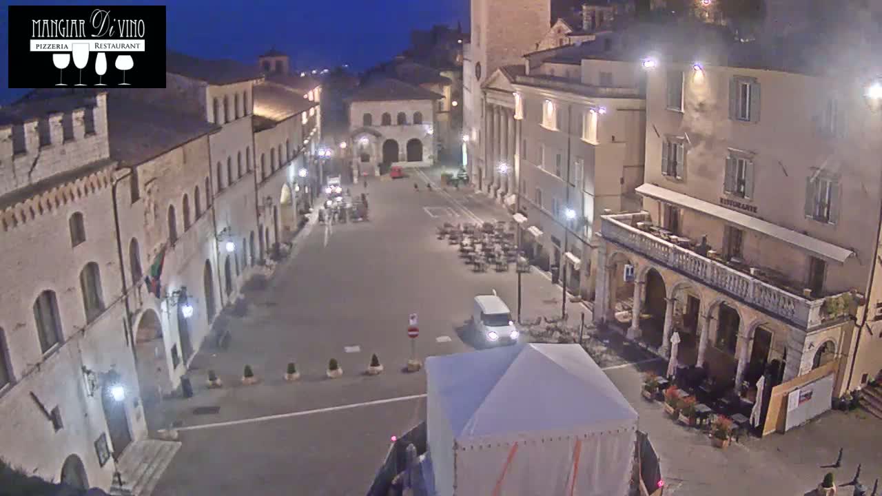 A mostly empty town square at night, lit by streetlights, is surrounded by stone buildings, with a few outdoor tables and chairs and a white van in the center.