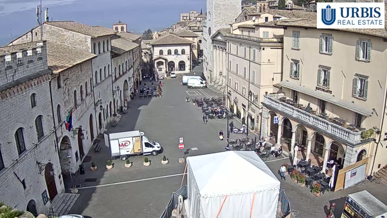 A town square in a historic Italian town on a sunny day, surrounded by stone buildings, with several people sitting at outdoor cafes and a white tent in the center.