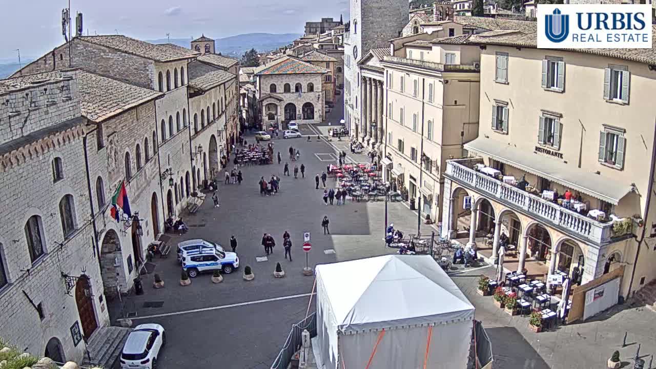 A sunny day reveals a town square surrounded by historical buildings, with people milling about and outdoor seating areas at cafes.