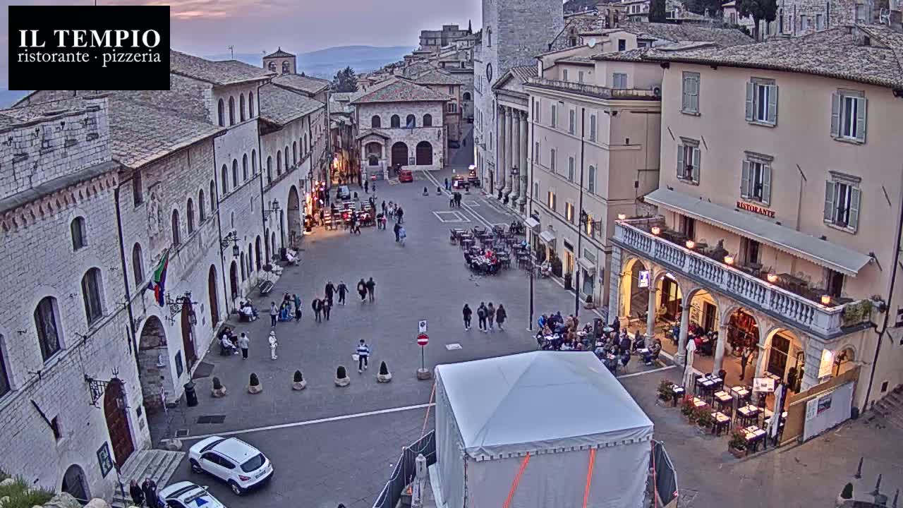 A large town square, surrounded by old stone buildings, is populated with people and outdoor seating at restaurants under a twilight sky.