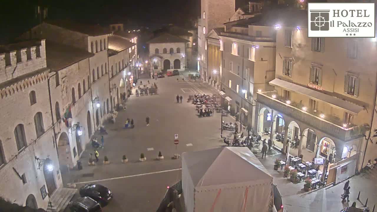 A nighttime, high-angle view shows a town square with several buildings surrounding it, some with outdoor seating areas, and a few people walking around.