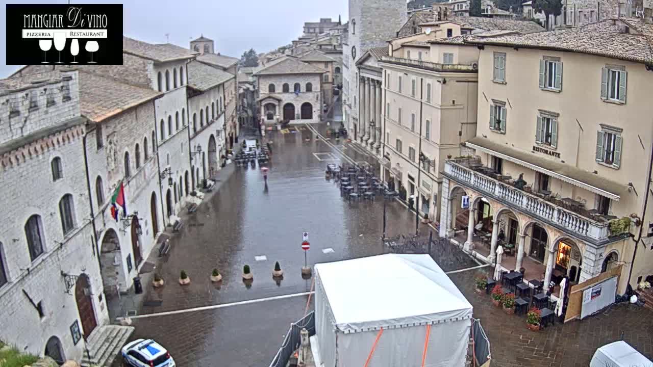 A rain-soaked town square in a European-style town, surrounded by old buildings, with a few cars and outdoor seating areas visible.