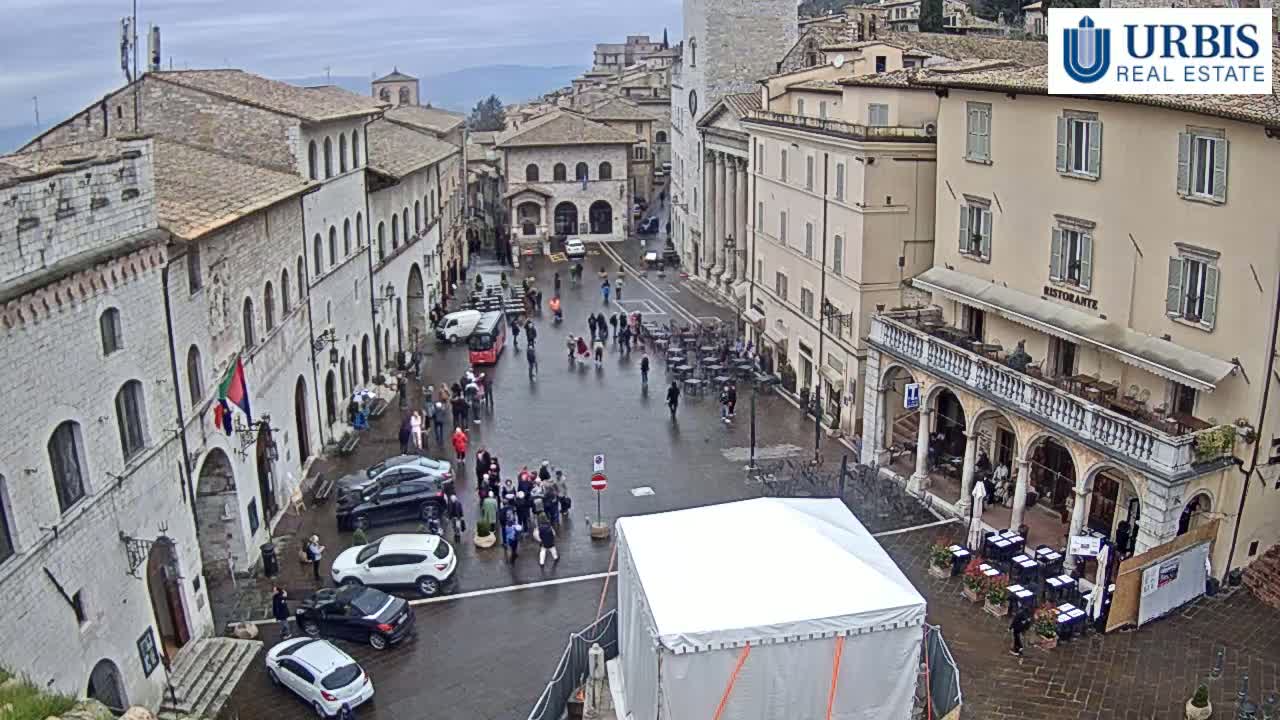 A wet town square in Europe, surrounded by historic buildings, with several people and parked cars, under an overcast sky.