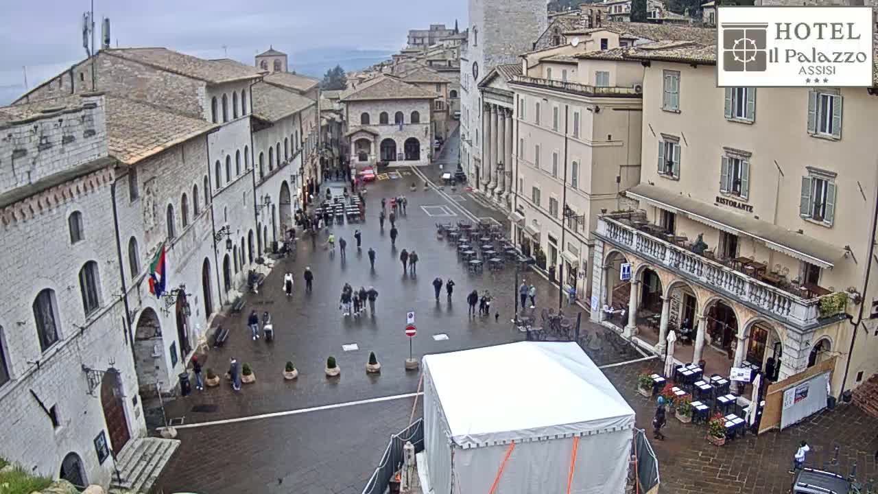 A wet, stone plaza in a European town is surrounded by multi-story buildings, with several people walking around and some outdoor seating.