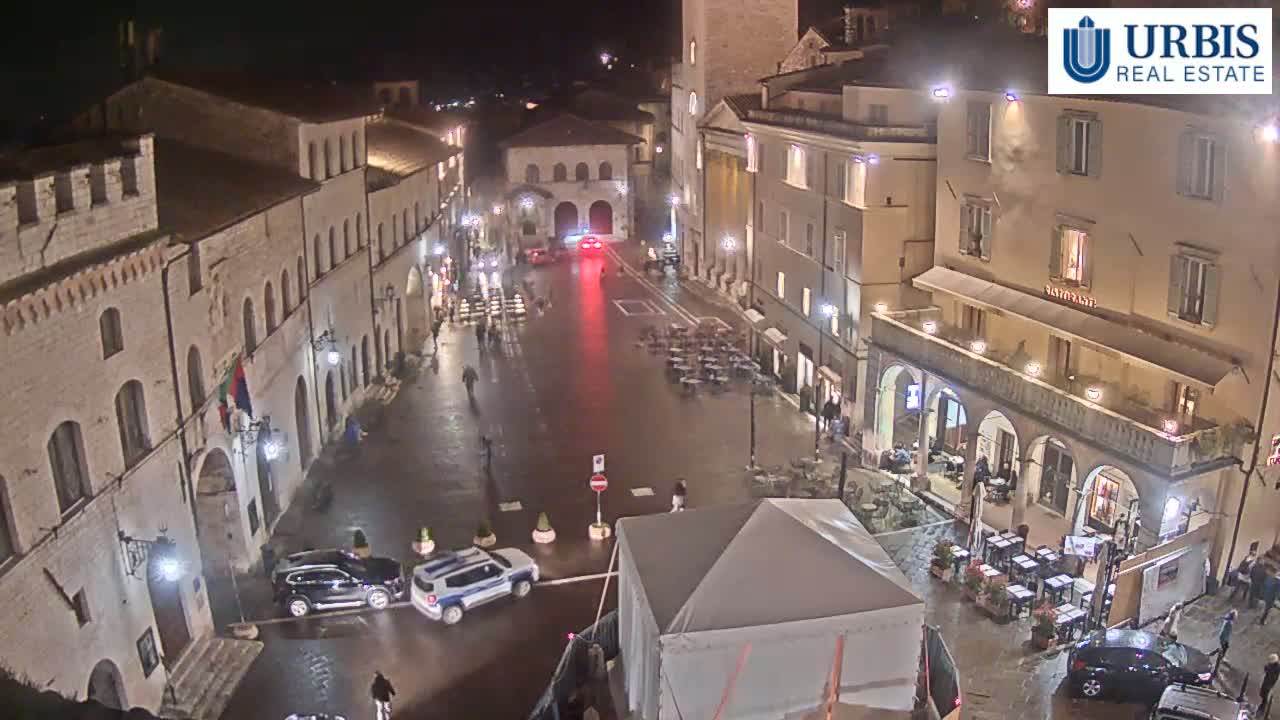 A wet, nighttime view of a town square in a European-style city, with several cars parked and a few people walking around.