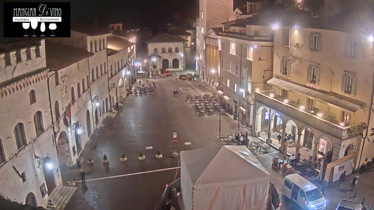 A wet, nighttime view of a town square surrounded by old buildings, with sparsely populated outdoor seating areas and a white van parked near a tent.