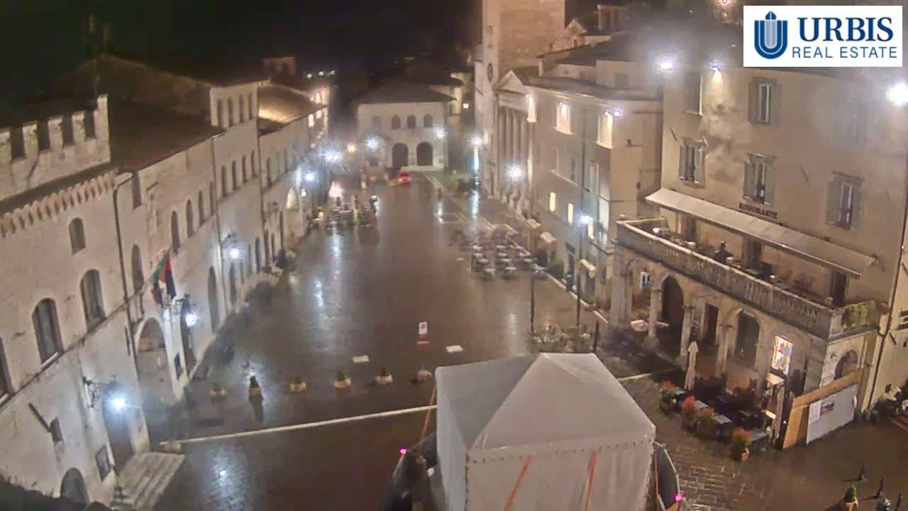 A rain-slicked town square at night, surrounded by historic-looking buildings, with a few small outdoor seating areas and a large tent in the center.