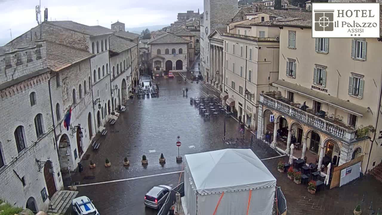 A rain-soaked town square is surrounded by aged stone buildings, with a few cars and a temporary tent visible.