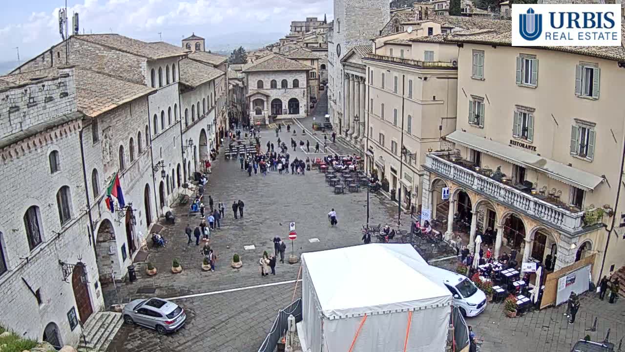 A large, stone plaza in a historic town is populated with people sitting at outdoor cafes and walking around under an overcast sky.