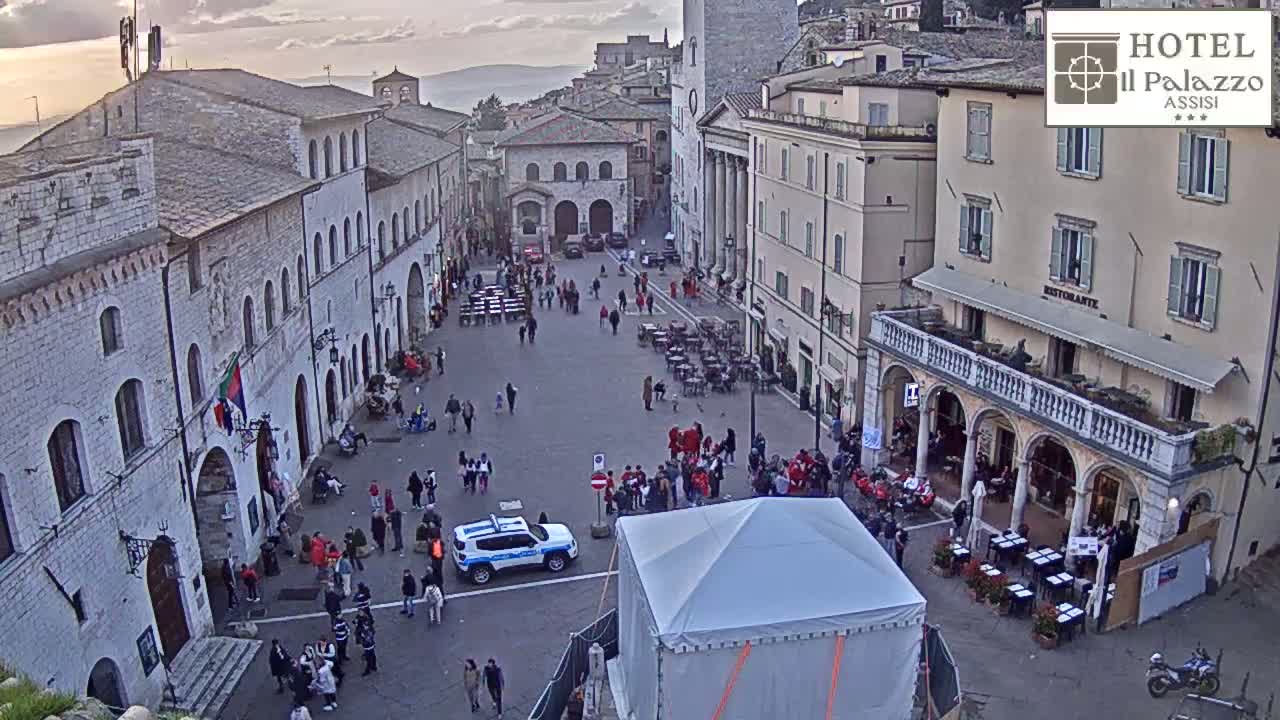 A town square in what appears to be a European town, with several stone buildings surrounding it, many people walking around, and a police car parked near a white tent, under overcast skies.