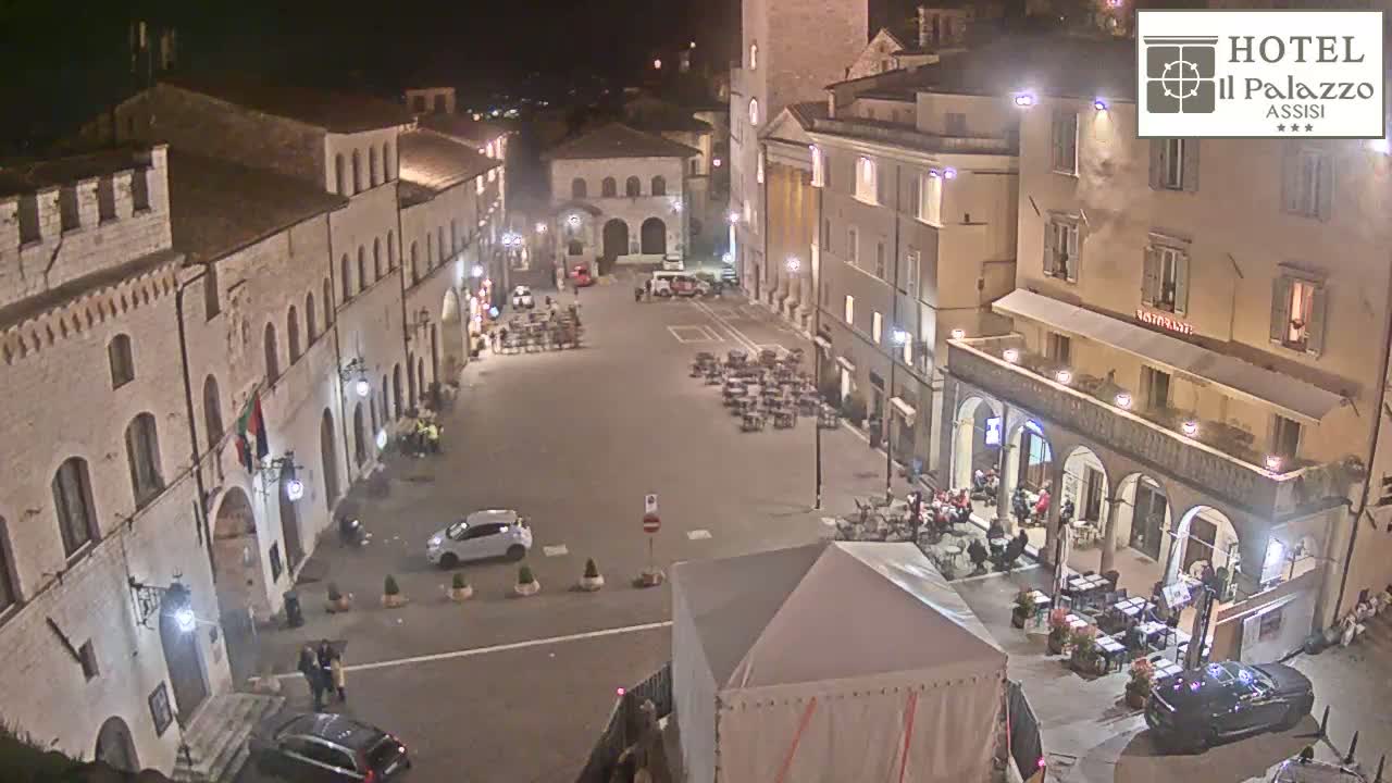 A nighttime, high-angle view of a town square with several buildings, parked cars, and outdoor seating areas at restaurants.
