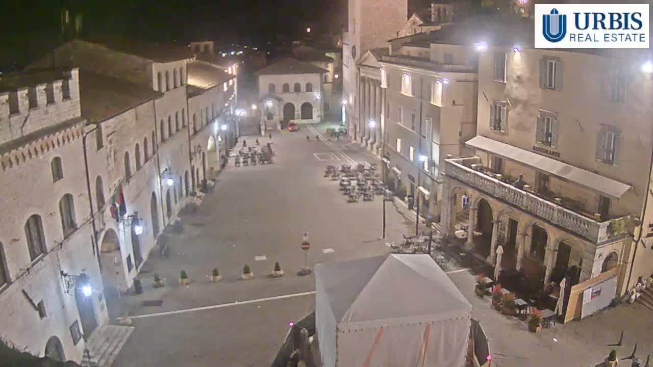 A mostly empty town square at night, illuminated by streetlights, is surrounded by historic-looking buildings.