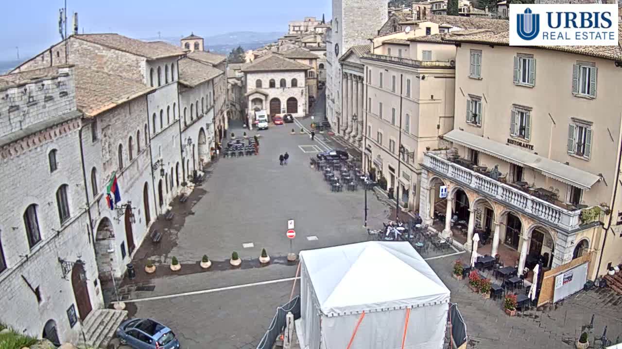 A large town square, mostly empty, is surrounded by ancient-looking stone buildings under a cloudy sky.