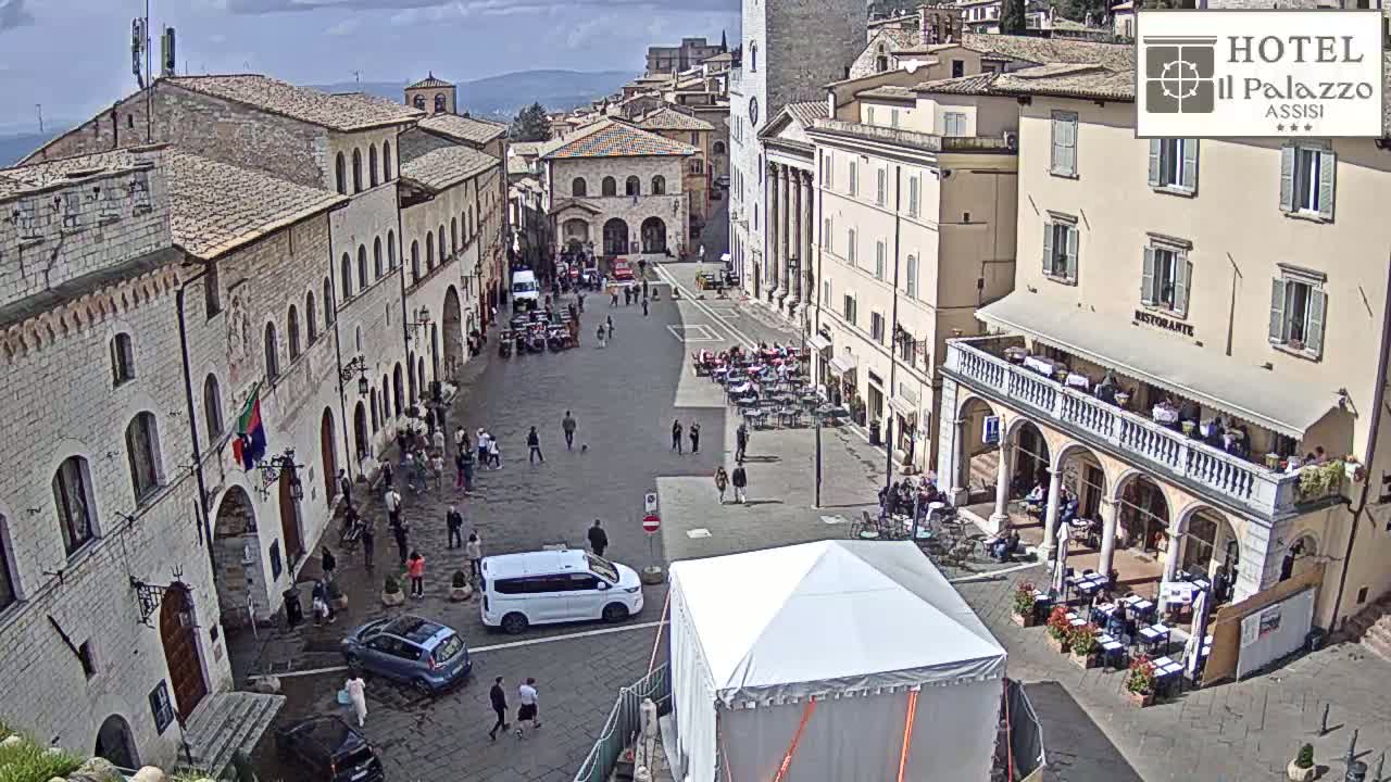 A partly cloudy day reveals a large town square in Italy, populated with people, vehicles, and outdoor seating areas in front of historic buildings.