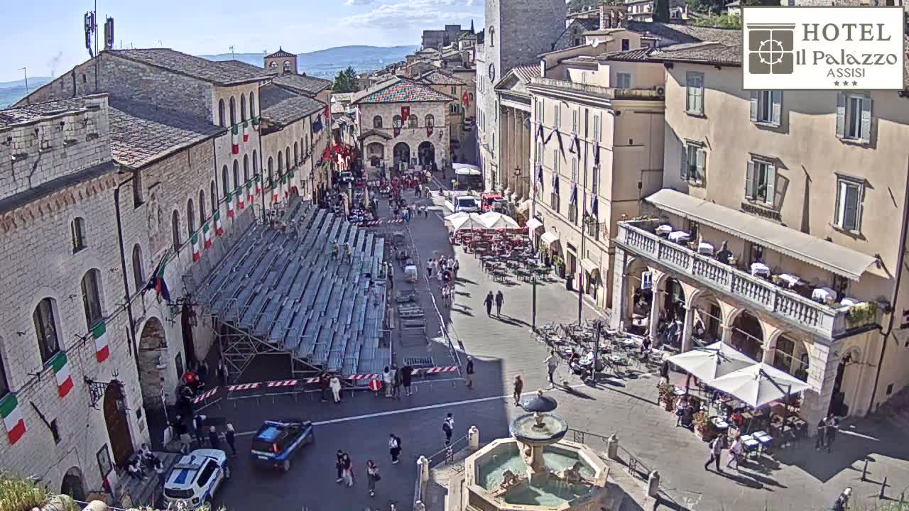 A sunny, outdoor view of a town square in Italy, showing a large crowd of people gathered around a temporary seating structure, buildings with Italian flags, and a fountain.